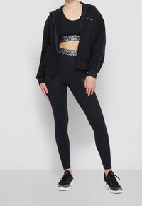Schwarzes Sportoutfit mit einem geschnittenen Tanktop, hochtaillierten Leggings und einem Reißverschluss-Hoodie. Alle Teile zeigen das Calvin Klein-Logo.