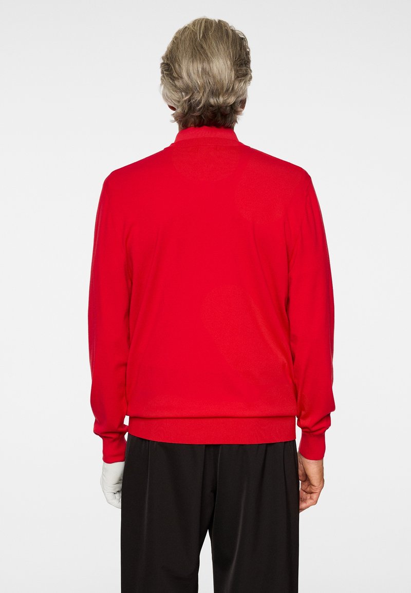 Person mit kurzen hellbraunen Haaren, trägt ein leuchtend rotes Langarmshirt und schwarze Hose, steht mit dem Rücken vor weißem Hintergrund.