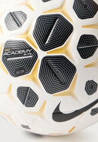 Piłka nożna Nike Academy Elite o teksturowanej białej powierzchni. Posiada czarne sześciokątne panele ze złotymi akcentami oraz wyraźne czarne logo Swoosh.