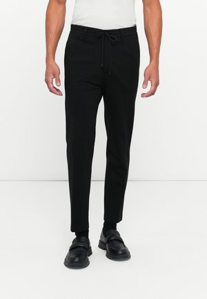 Black tapered pants with an elastic waistband and drawstring, crafted from a smooth fabric. Worn with black loafers and ankle socks.