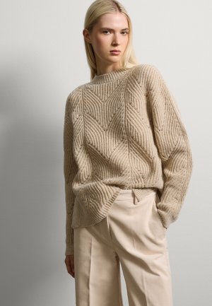 Maglione beige lavorato a maglia con una texture a coste e motivo a zigzag, caratterizzato da un collo rotondo e una vestibilità ampia, abbinato a pantaloni beige chiaro.