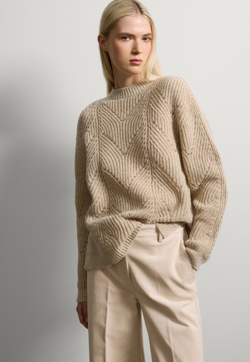 Beige Strickpullover mit gerippter Textur und Zickzackmuster, runder Ausschnitt und lockerer Passform, kombiniert mit hellbeigen Hosen.
