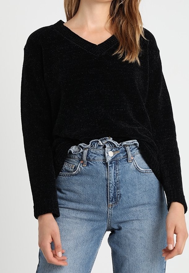 Černý, strukturovaný svetr s dlouhým rukávem a výstřihem do V ve spojení s vysoce pasovanými světle modrými džíny s volánkovým pasem a předním zapínáním na knoflíky.