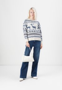 Strickpullover in Creme mit blauen Rentieren und geometrischem Muster, kombiniert mit dunkelblauen Schlagjeans und einer weißen Handtasche mit Spitzendetails.
