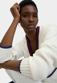 Maglione bianco a maglia con accenti blu navy e rossi sul collo e sui polsini; presenta un profondo scollo a V e dettagli a coste sulle maniche.