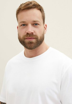 Bärtiger Mann mit kurzem hellbraunem Haar, blauen Augen, der ein einfaches weißes T-Shirt trägt und ruhig in die Kamera vor hellem Hintergrund blickt.