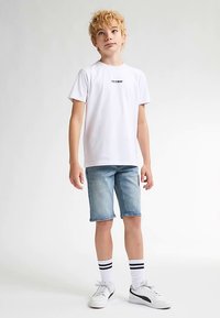 Camiseta blanca de manga corta con logo estampado, combinada con shorts de mezclilla azul claro. Completado con zapatillas blancas y calcetines a rayas.