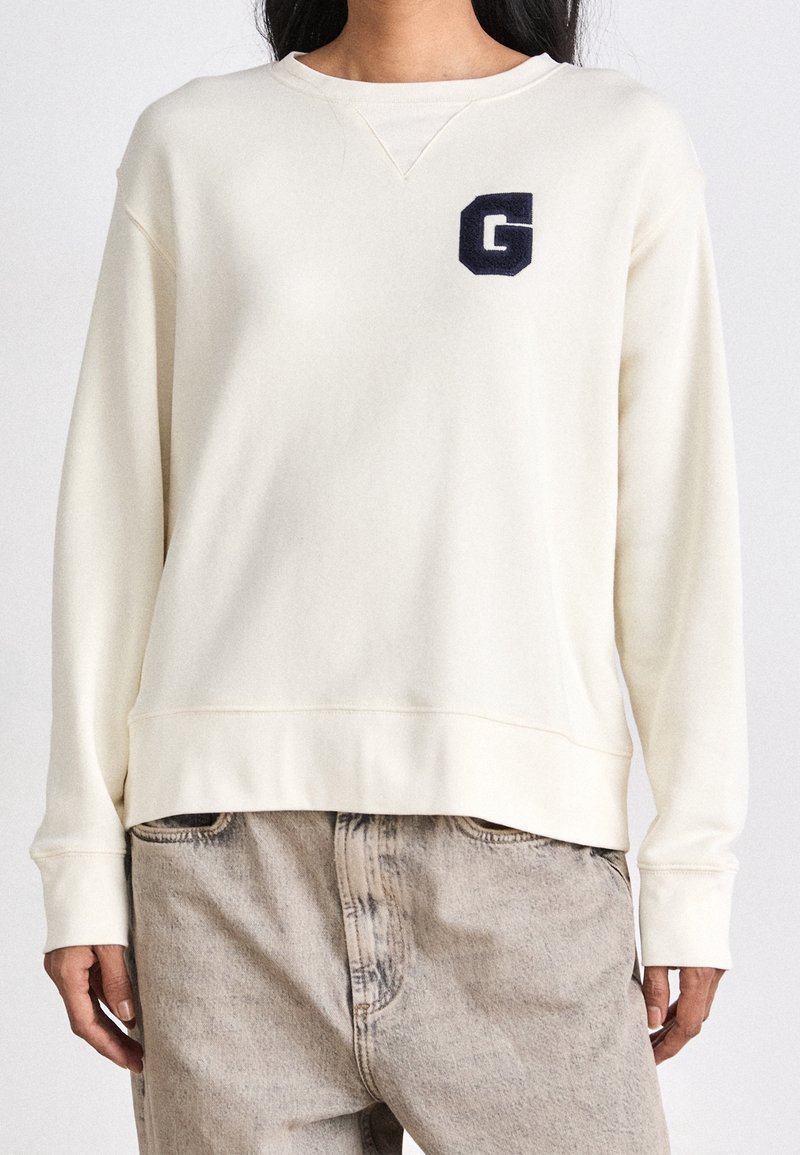 Femme portant un sweat-shirt crème à manches longues avec un logo sombre "G" sur la poitrine et un pantalon beige clair décontracté.