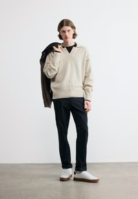 Νεαρός άνδρας στέκεται κρατώντας ένα σκοτεινό σακάκι στον ώμο του, φοράει μπεζ πουλόβερ, μαύρο παντελόνι και λευκά αθλητικά παπούτσια σε γκρι πάτωμα.