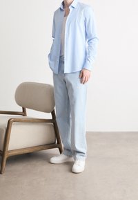 Lichtblauw overhemd met knopen, grijze tanktop, losse lichtblauwe jeans, witte sneakers, staande naast een beige gestoffeerde stoel met houten frame.