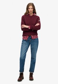 Bordeaux gebreide trui over een rood-wit geruit shirt, gecombineerd met blauwe opgerolde jeans en bruine veterschoenen.