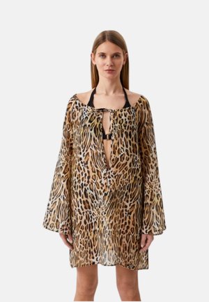 Donna che indossa un abito largo a maniche lunghe con stampa leopardata sopra un top nero con scollo halter, in piedi contro uno sfondo chiaro e semplice.