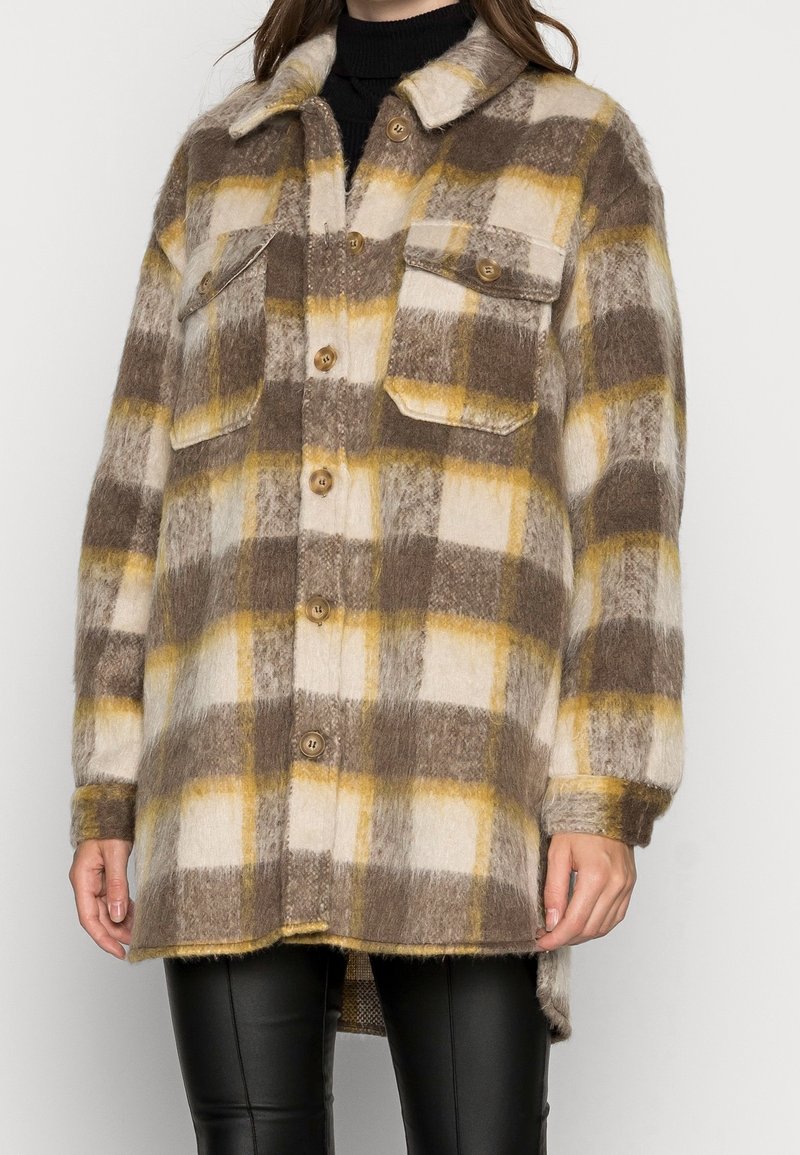 Femme portant une veste en peluche à carreaux marron, crème et jaune avec des poches poitrine et un pantalon noir.