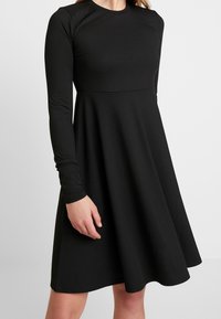 Musta ribbed pitkähihaisista mekosta, jossa on tiukka yläosa ja leveä helma. Sujuva rakenne, pyöreä kaula-aukko ja hienovaraiset saumarivistöt vyötäröllä.