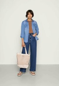 Donna in piedi che indossa una camicia di jeans blu, un top marrone, jeans a vita alta, sandali, e tiene una borsa a tracolla beige a righe al suo fianco.