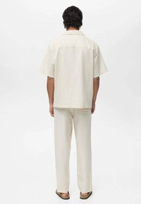 Cămașă scurtă de culoare bej deschis, cu croială dreaptă, asortată cu pantaloni drepți din același material, ambele dintr-un țesătură ușoară.