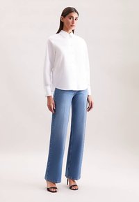 Biała koszula na guziki z długimi rękawami, zestawiona z jasnoniebieskimi szerokimi dżinsami. Całość uzupełniają czarne sandały na obcasie.