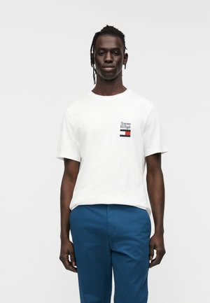 Jeune homme aux cheveux tressés portant un t-shirt blanc Tommy Hilfiger et un pantalon bleu, debout devant un fond clair uni.