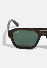 Ochelari de soare în stil broască țestoasă cu ramă pătrată, lentile verzi, accente argintii și logo-ul Ray-Ban pe partea dreaptă din față.