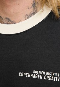 Camiseta de algodón negra con un cuello redondo de canalé crema, que presenta un texto blanco que dice "HOLMEN DISTRICT COPENHAGUE CREATIVO" en el frontal.