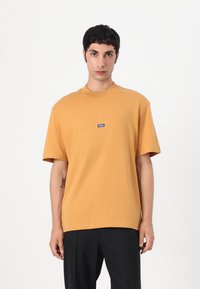 Kortærmet, sennepsgul T-shirt lavet af bomuld, med en rund halsudskæring og et lille blåt logo-patch på brystet.