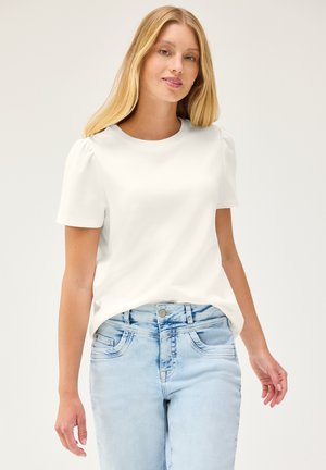 Joven mujer con cabello rubio largo que lleva una camiseta blanca de manga corta y vaqueros de mezclilla azul claro, de pie frente a un fondo sencillo.