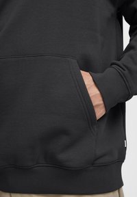 Sudadera negra con una textura suave, un bolsillo tipo canguro, puños y dobladillo acanalados. Presenta una pequeña etiqueta en la parte inferior derecha.