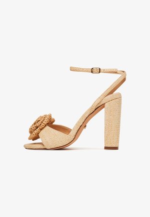 Beżowy pleciony sandał na wysokim obcasie typu blok, z odkrytym noskiem i warkoczowym kwiatowym detalem na przednim pasku. Regulowany pasek wokół kostki.