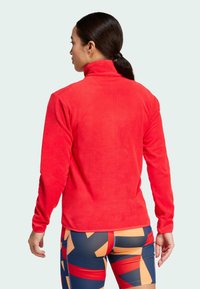 Roter Fleece-Pullover mit hohem Kragen, langen Ärmeln und glatter Textur. Besitzt ein tailliertes Design, kombiniert mit mehrfarbigen gemusterten Leggings.