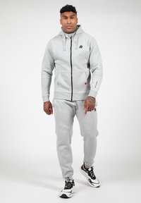 Grijze rits hoodie met kangoeroe pockets, gecombineerd met bijpassende sweatpants. Heeft een logo en een contrasterende zwarte rits. G gedragen met zwart-witte sneakers.