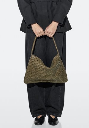 Osoba v černém oblečení držící velkou olivově zelenou pletenou kabelku s pleteným uchem před nohama.