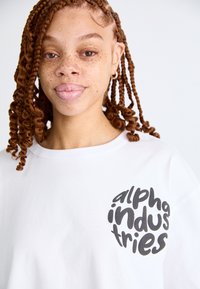 Λευκό βαμβακερό μπλουζάκι με κοντά μανίκια, με μια μεγάλη μαύρη γραφιστική εκτύπωση που γράφει "alpha industries" μπροστά.