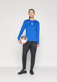 Blue long-sleeve athletic shirt with logo, paired with black athletic pants and black soccer shoes. Holding a patterned soccer ball.
