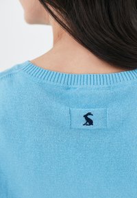 Kobieta w jasnoniebieskim swetrze z małym czarnym logo królika na plecach w pobliżu dekoltu, z ciemnobrązowymi włosami opadającymi na ramię.