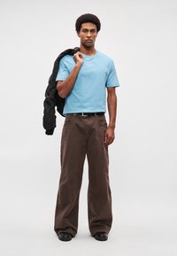 Helehall sinine lühikeste varrukatega särk logoga, koos lahtiste pruunide pükstega ja mustade kingadega, hoides õlal musta jopet.