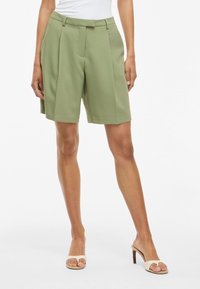 Pantalones cortos verdes a medida en tejido ligero, con pliegues, bolsillos laterales y dobladillo vuelto. Combinados con tacones abiertos color piel.