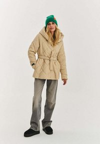 Béžová prošívaná bunda s odnímatelnou kapucí, pásem na zavázání a bočními kapsami, v kombinaci se šedými rozšířenými džíny a černými nazouvacími botami. Zelená pletená čepice.