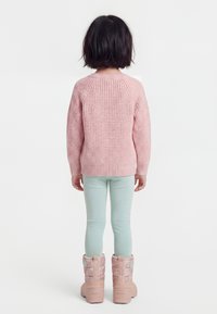 Dítě s krátkými tmavými vlasy, oblečené v růžovém pleteném svetru, světle modrých kalhotách a růžových zimních botách, stojící zády k divákovi na bílém pozadí.