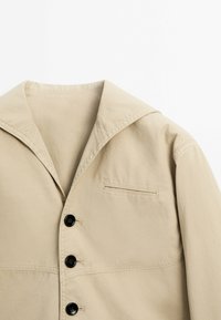 Beige jacket with wide collar, three black buttons, and a stitched welt pocket on the chest, shown against a white background.