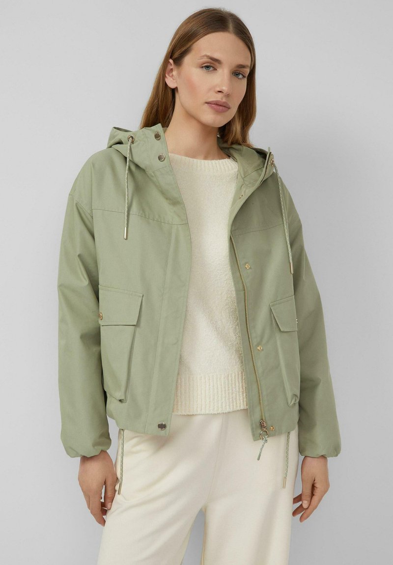 Kobieta w jasnozielonej kurtce z kapturem na kremowym swetrze i białych spodniach, stojąca na tle jednolitego szarego tła.