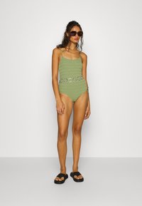 Maillot de bain une pièce vert olive avec une texture en zigzag, des bretelles fines et une ceinture ronde argentée ; associé à des tongs noires.