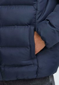 Marineblå puffyjakke med en quiltet design, som har en sidelomme og en glatt, blank stofftekstur.