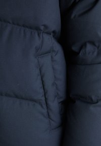 Marinblå vadderad jacka med en sidoficka. Materialet ser mjukt ut med en slät yta och puffig stoppning.