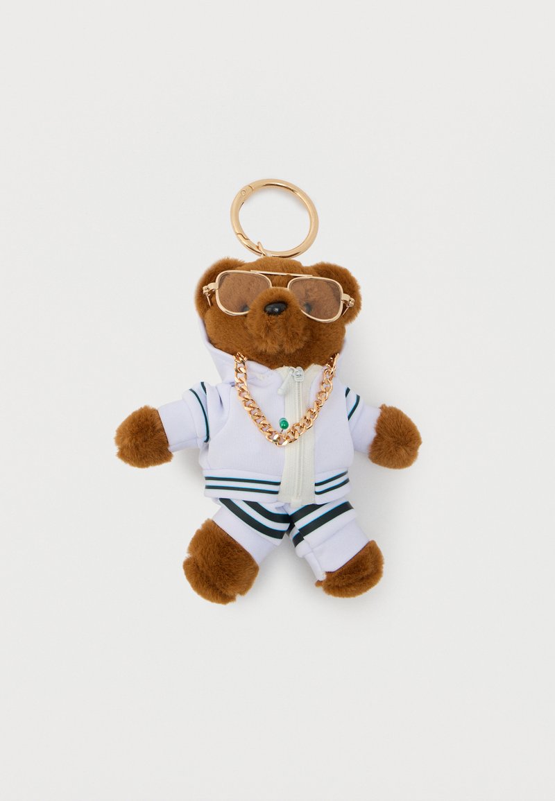 Porte-clés petit ours en peluche marron portant un survêtement blanc, un collier en chaîne dorée et des lunettes de soleil à monture dorée sur un fond uni.