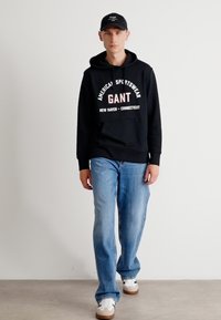 Čierna mikina s kapucňou a bielym nápisom "GANT AMERICAN SPORTSWEAR", v kombinácii so svetlomodrými džínsami a bielymi teniskami. Čierna čiapka.