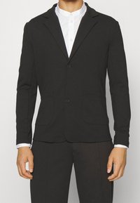 Sort blazer lavet af blødt stof, med to-knap lukning, notch kraver og to forlommer. Båret over en hvid skjorte med krave.