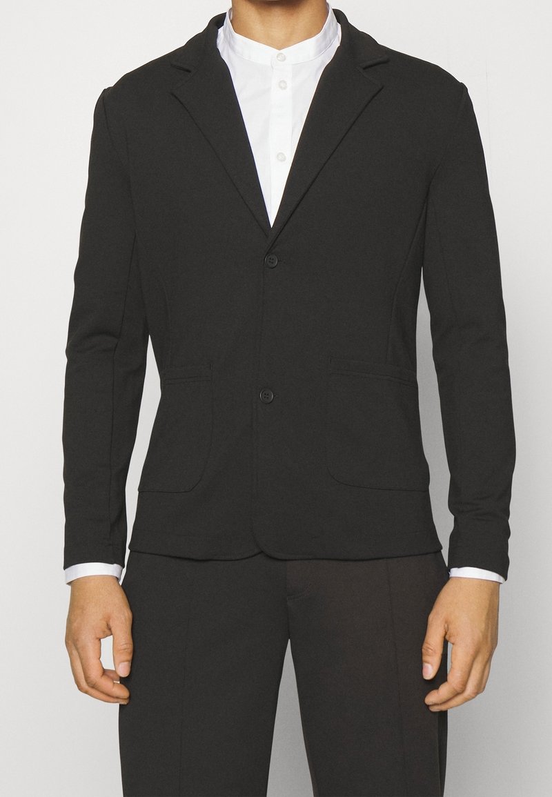 Schwarzer Blazer aus weichem Stoff, mit einer Zwei-Knopf-Verschluss, Reverskragen und zwei Fronttaschen. Getragen über einem weißen Hemd mit Kragen.