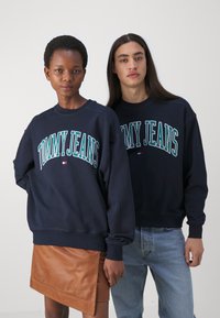 Sudaderas azul marino con el logo "TOMMY JEANS" en turquesa, cuello redondo y puños de canalé, llevadas por dos modelos con acentos de denim y cuero.