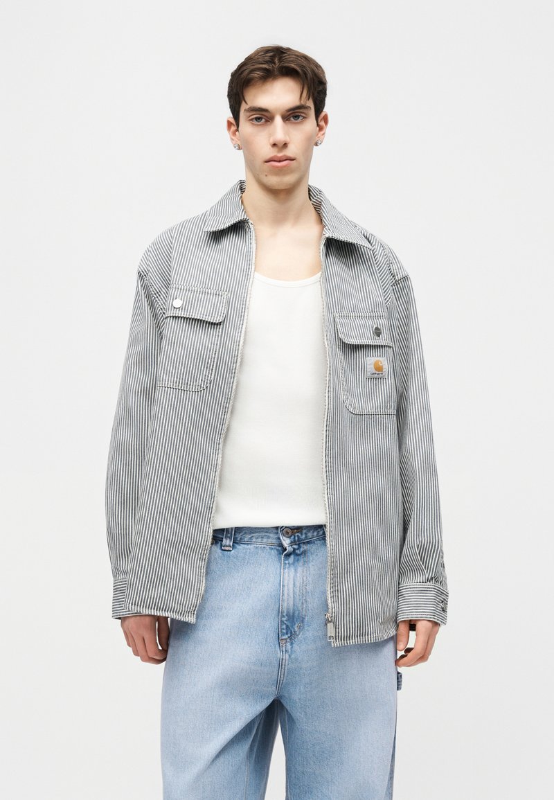 Giovane uomo con giacca grigia a righe con cerniera sopra una canottiera bianca e jeans azzurri, in piedi davanti a uno sfondo bianco uniforme.