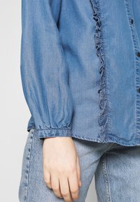 Denim shirt with ruffled front detailing, long sleeves with elastic cuffs, in light blue, featuring black buttons and a relaxed fit.